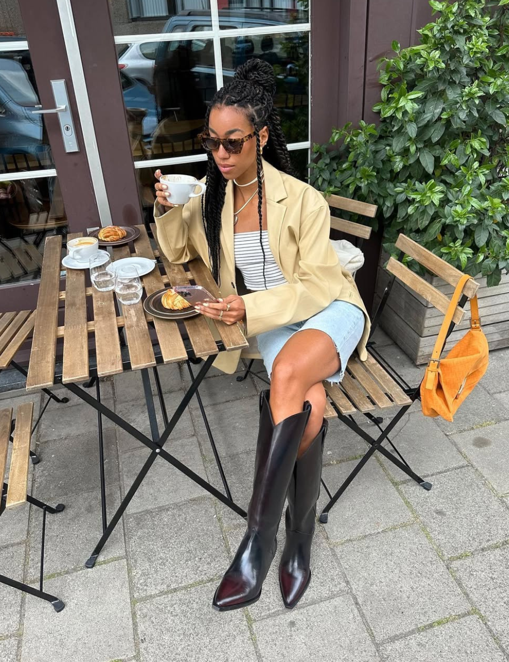 Modelo posa em café com bermuda jeans, bota longa preta, blazer bege e bolsa caramelo em clima urbano elegante.