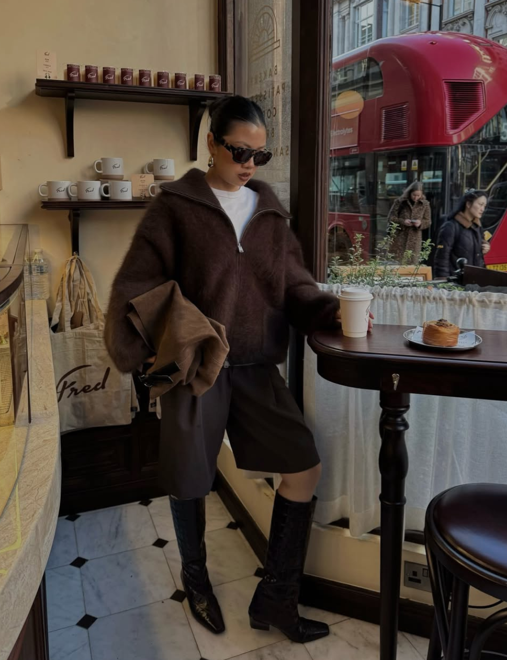 Modelo posa com bermuda e bota longa pretas, malha marrom ampla e óculos escuros em café elegante e urbano.