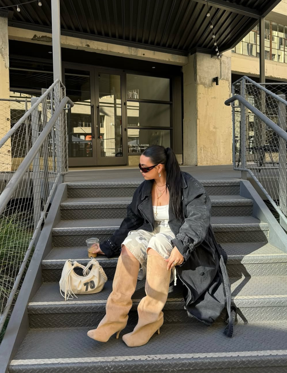 Modelo posa sentada na escada com bermuda clara, bota longa bege, casaco preto oversized e mood urbano sofisticado.