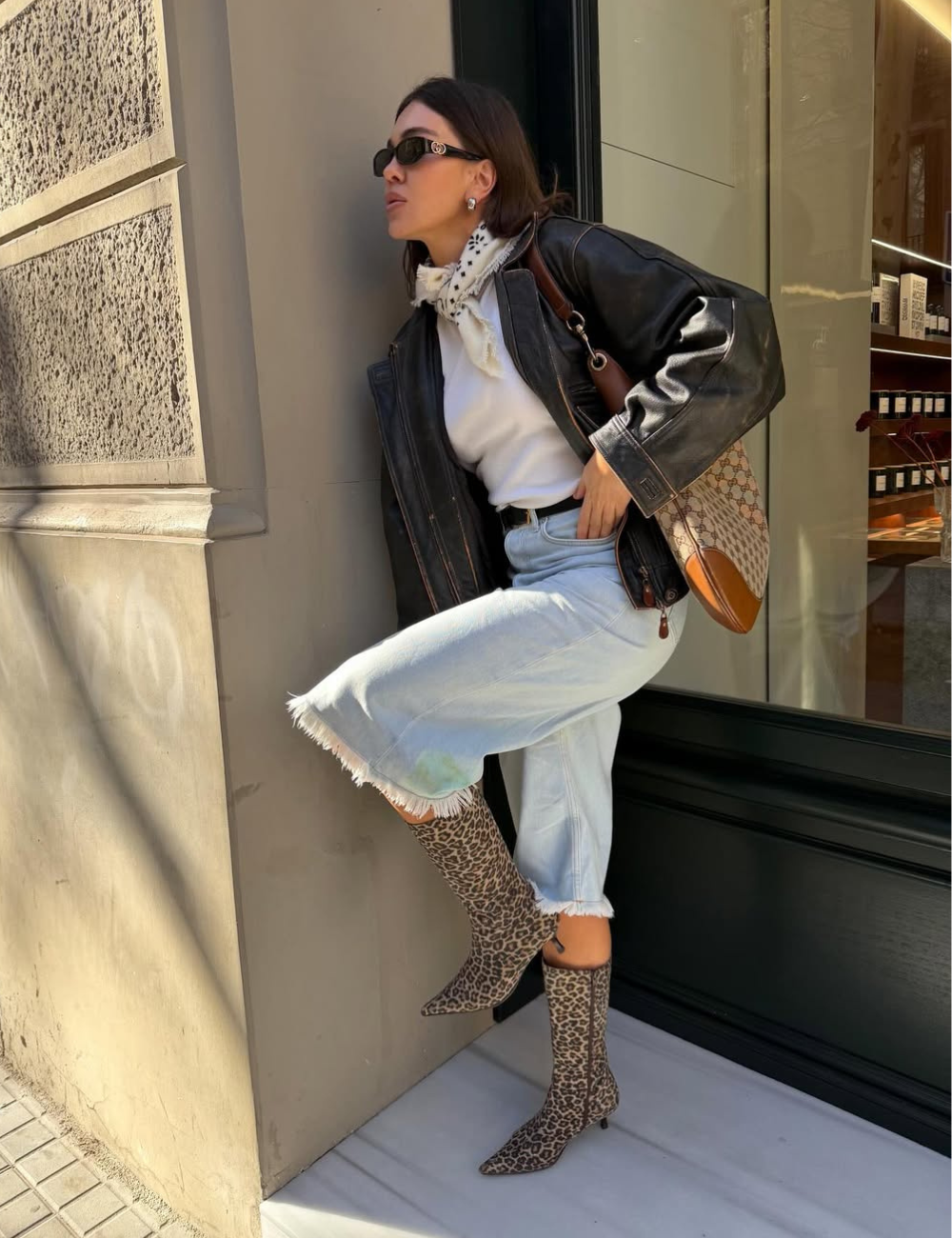 Modelo posa com bermuda jeans clara e bota longa animal print, jaqueta preta e lenço branco em clima urbano sofisticado.