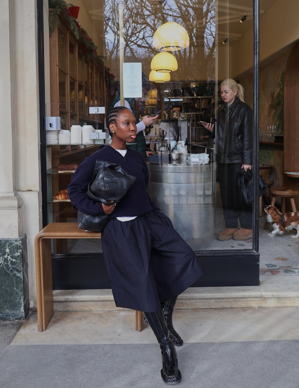 Modelo posa com bermuda ampla e bota longa preta, tricô escuro e bolsa de couro diante de café elegante e sereno.