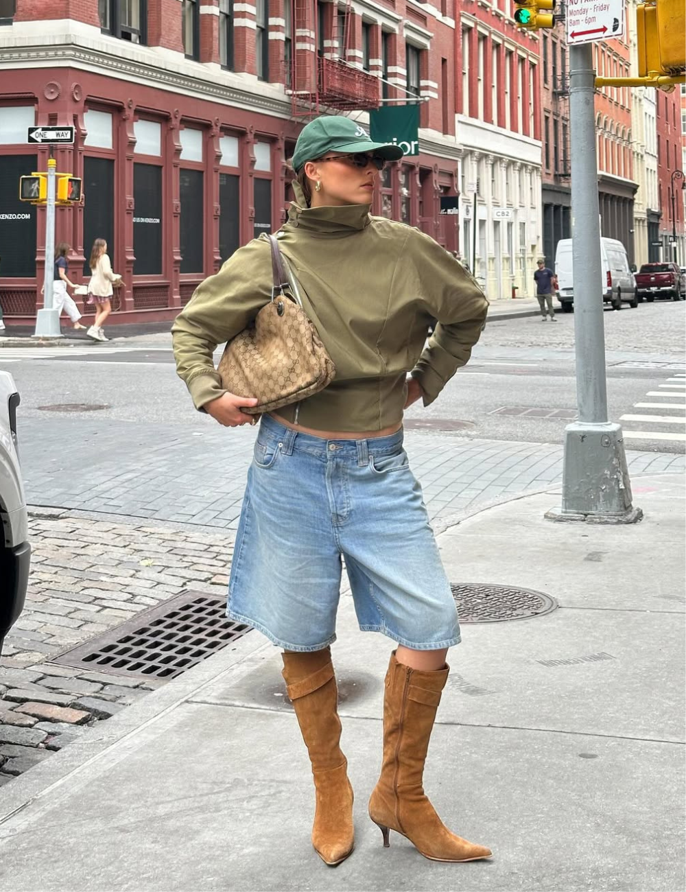 Modelo posa com bermuda jeans ampla e bota longa de camurça caramelo, jaqueta verde e boné em rua urbana elegante.