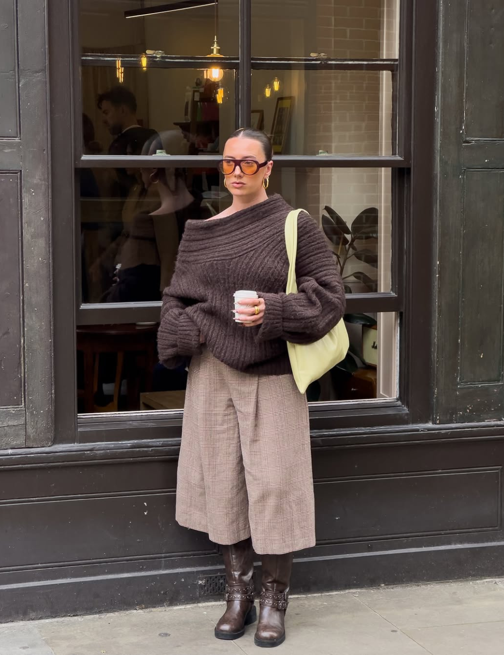 Modelo posa com bermuda xadrez e bota longa marrom, tricô amplo e bolsa clara diante de vitrine, em clima urbano sóbrio.