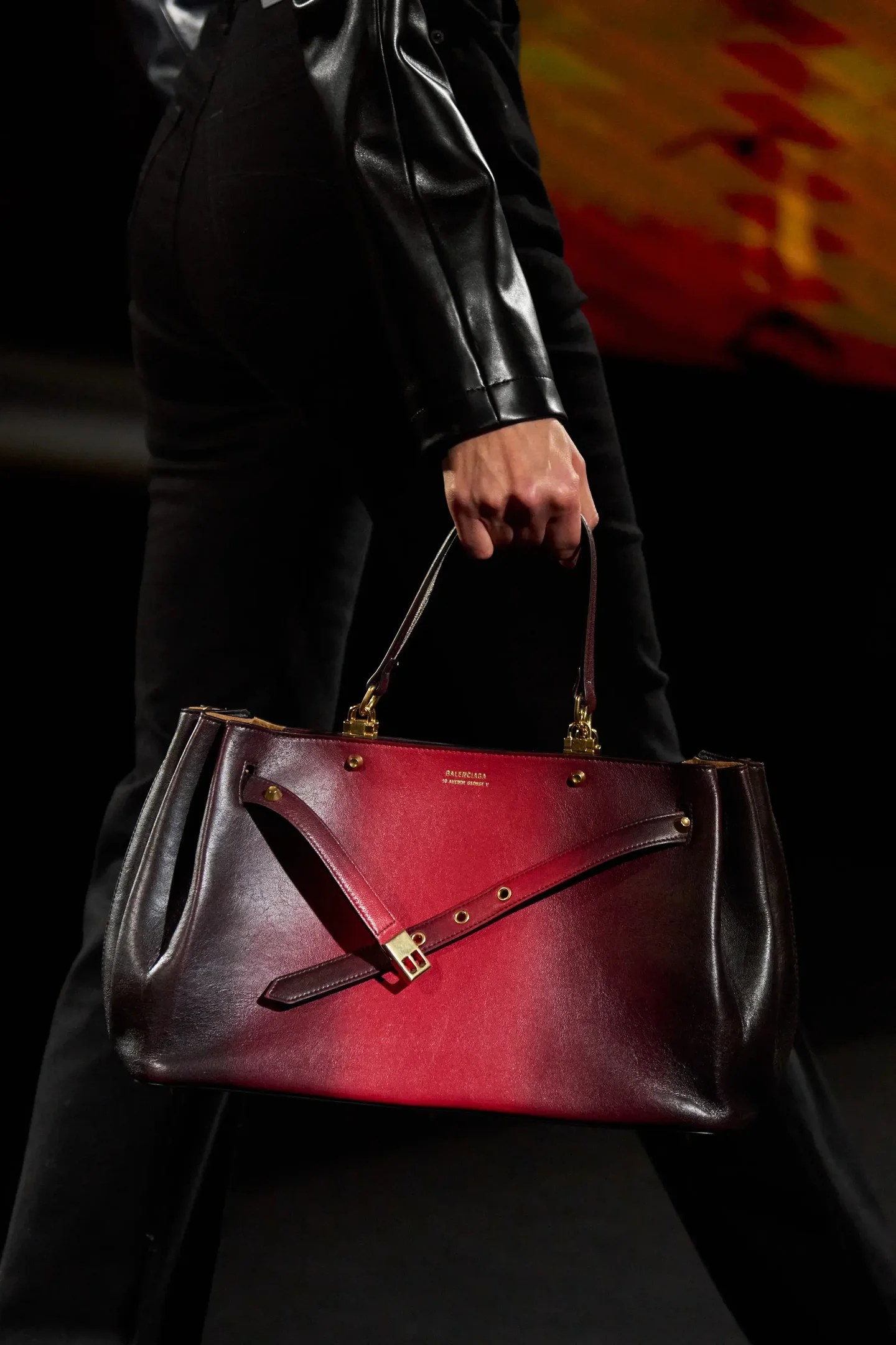 Modelo segura bolsa de couro vermelho e marrom com fivela, vestindo jaqueta e calça preta, estilo sofisticado para temporada