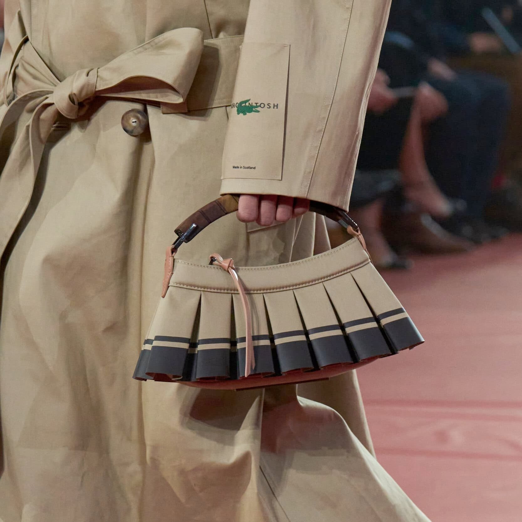 Trench coat bege com modelagem ampla e cinto estruturado em tecido resistente combina com bolsa plissada em tons neutros