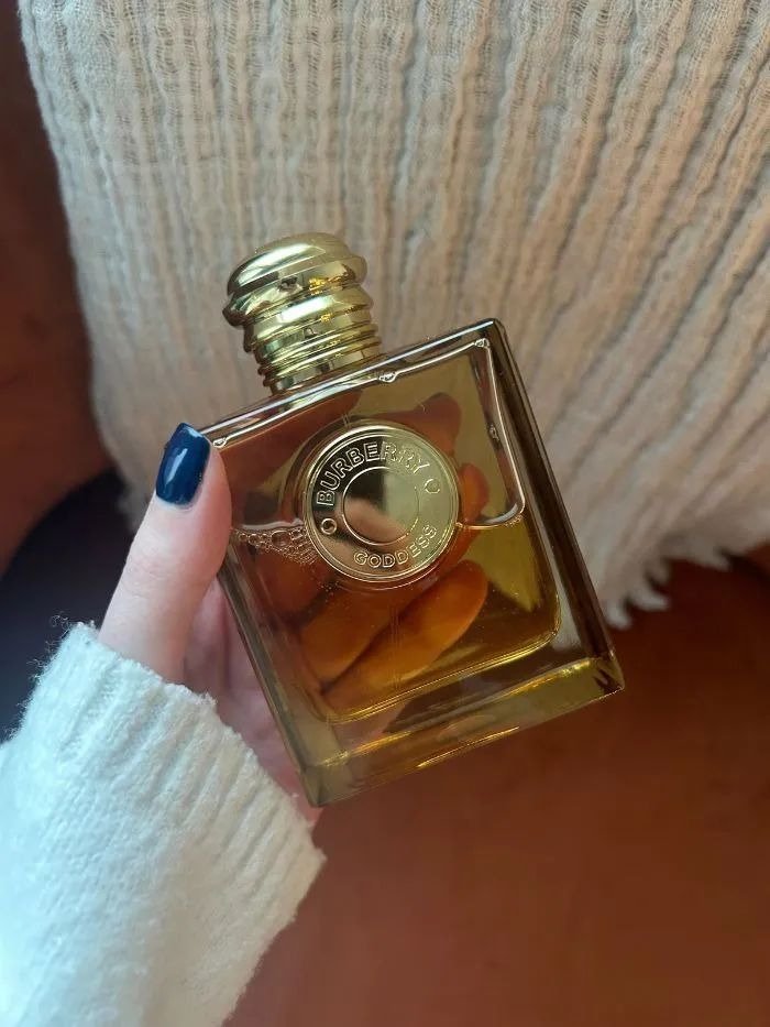 Frasco quadrado dourado do perfume Burberry Goddess, com tampa arredondada e detalhe circular em relevo, em mão com esmalte
