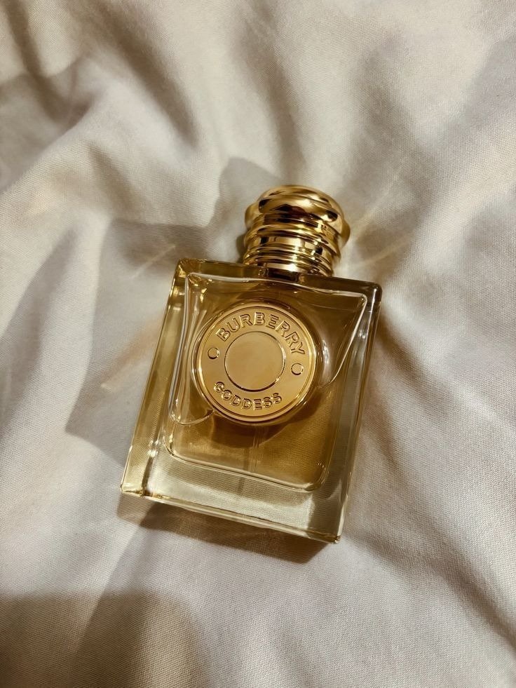 Frasco Burberry Goddess em vidro transparente com líquido dourado, tampa e detalhe frontal metálico dourado, sobre tecido