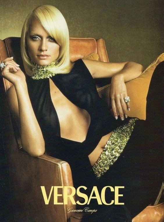 Modelo posa com vestido preto Versace de decote assimétrico e detalhes dourados, em clima sofisticado e elegante.