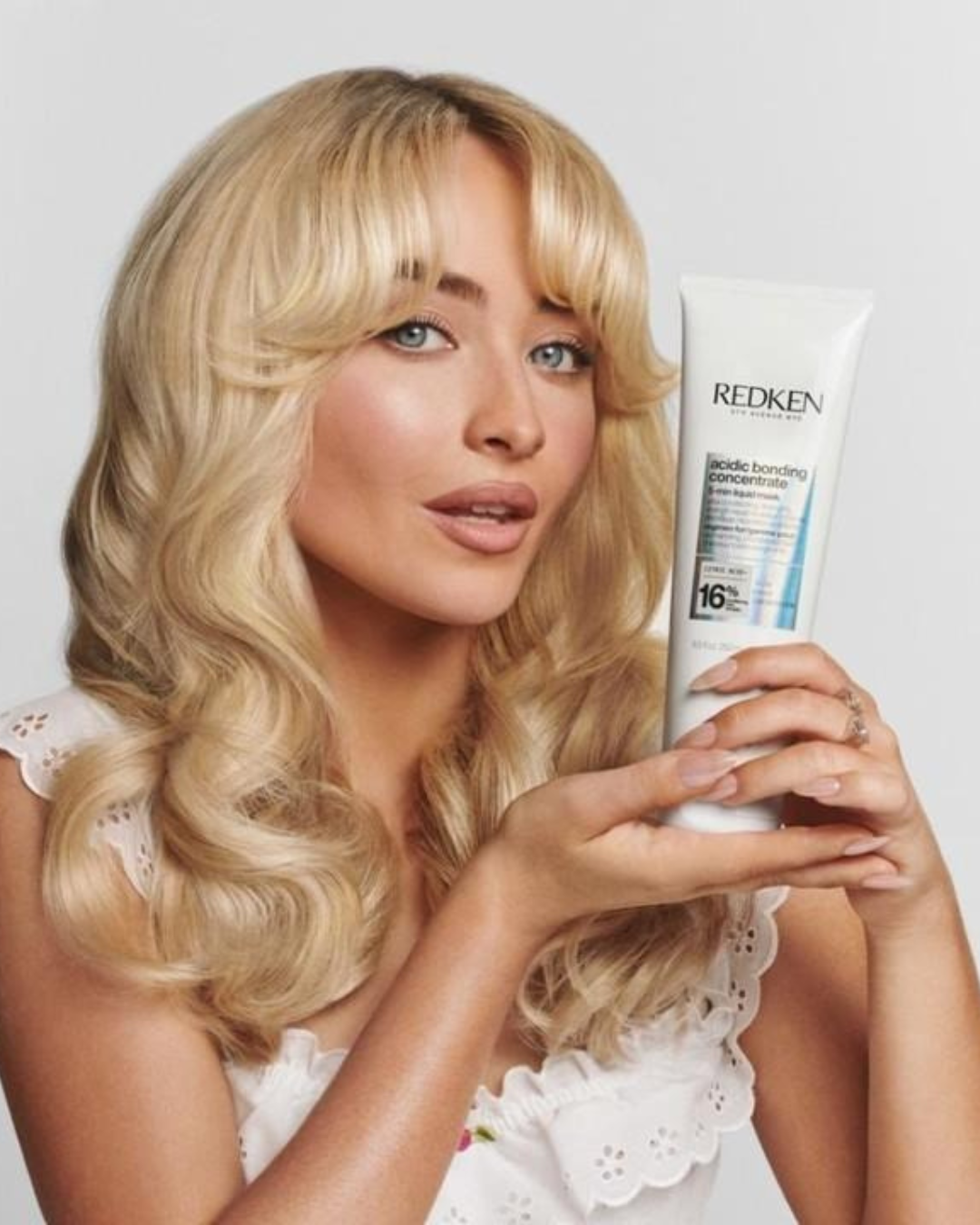 Cabelo da Sabrina Carpenter loiro ondulado com franja moldura o rosto; ela posa de branco com REDKEN. Texto: REDKEN 16%