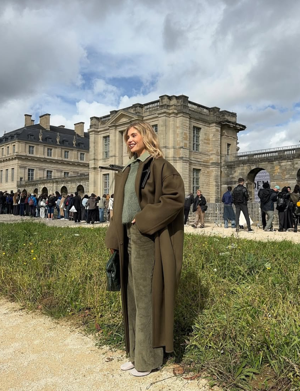 Mulher em pé em ambiente externo durante o dia. Usa calça de veludo verde, casaco longo e segura bolsa verde.
