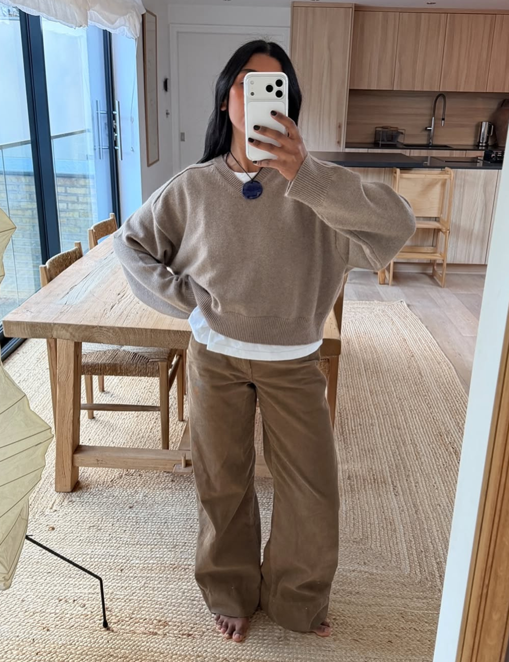 Mulher tira foto no espelho usando calça de veludo marrom, suéter bege e blusa branca por baixo.