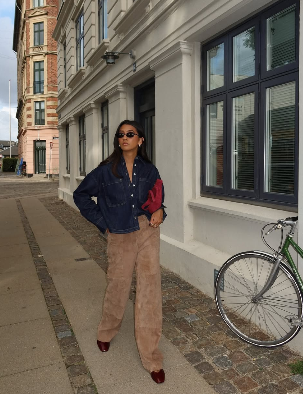 Mulher em pé na rua usando calça de veludo cor caramelo, camisa azul-escuro e sapatos fechados. Segura bolsa vermelha.