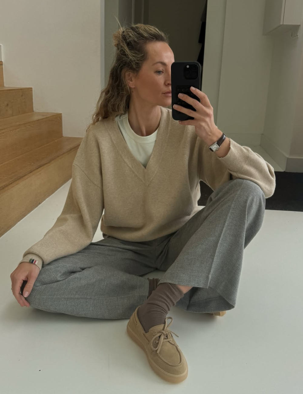 Modelo posa sentada com calças confortáveis para trabalhar em cinza, tricô bege e tênis neutro, em clima sereno.