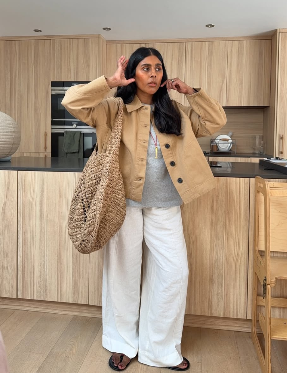 Modelo posa com calças confortáveis para trabalhar, jaqueta bege e tricô cinza em cozinha clara, com ar casual elegante.