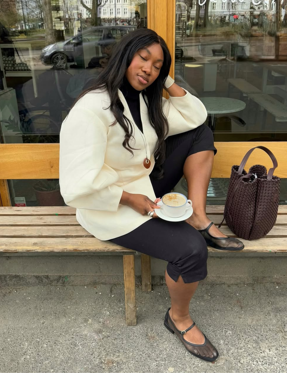 Modelo posa com calças confortáveis para trabalhar, blazer creme e sapatilha preta em clima sereno de café urbano.