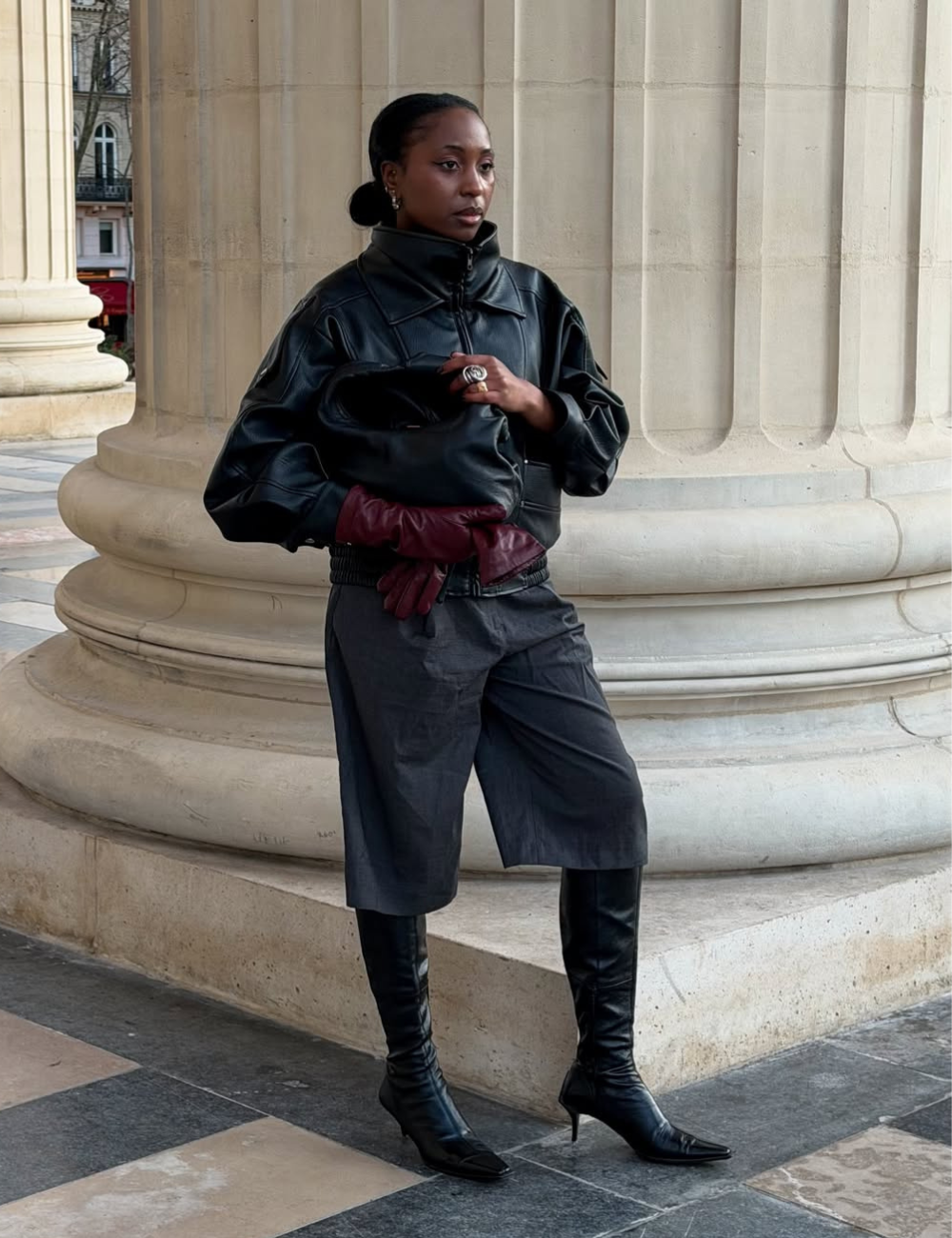 Modelo usa casaco de couro preto com gola alta, luvas bordô, bermuda de alfaiataria e botas de cano longo.
