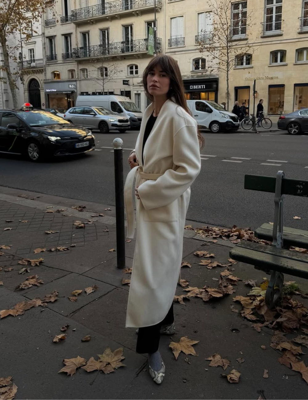 Mulher em pé usa um dos casacos femininos tendência na cor off-white enquanto está de pé em meio as folhas.