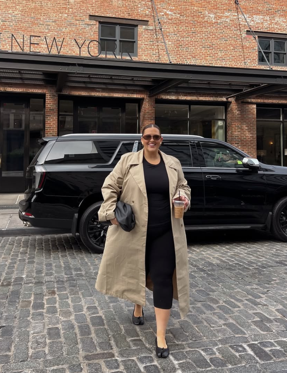 Modelo caminha em pé sorrindo enquanto segura um café. Usa trench coat bege, calça capri preta e sapatilhas.