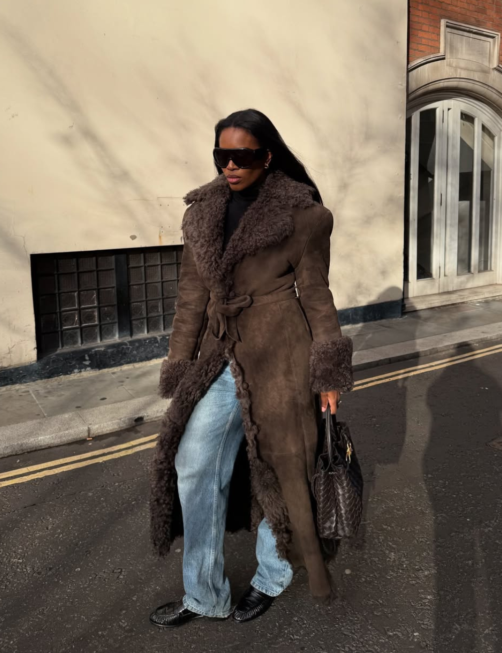 Mulher anda na rua usando óculos de sol, casaco teddy estilo penny lane, jeans claros e botas pretas.