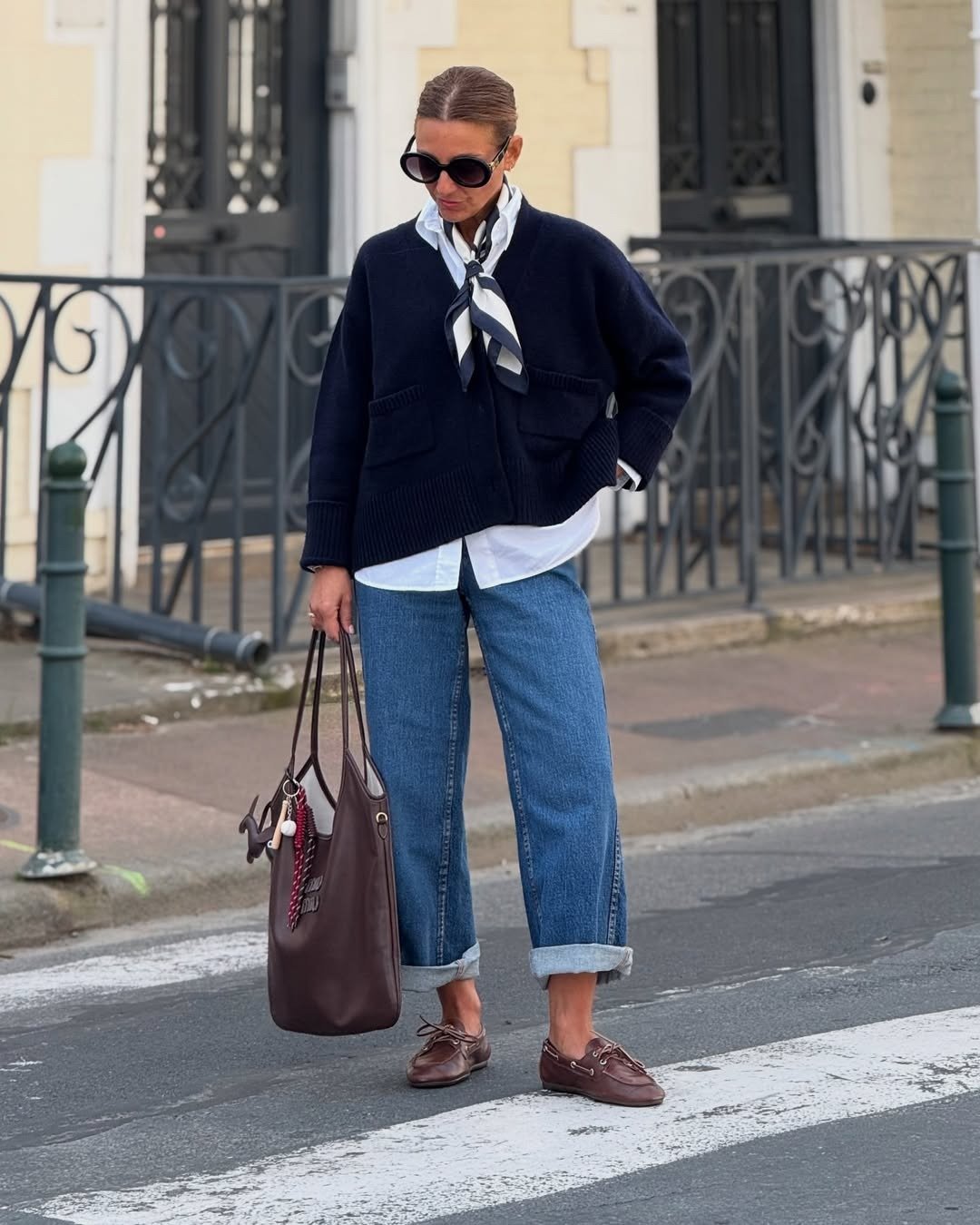 Mocassim feminino com calça jeans reta dobrada, camisa branca, tricô azul-marinho e lenço criam look urbano elegante.
