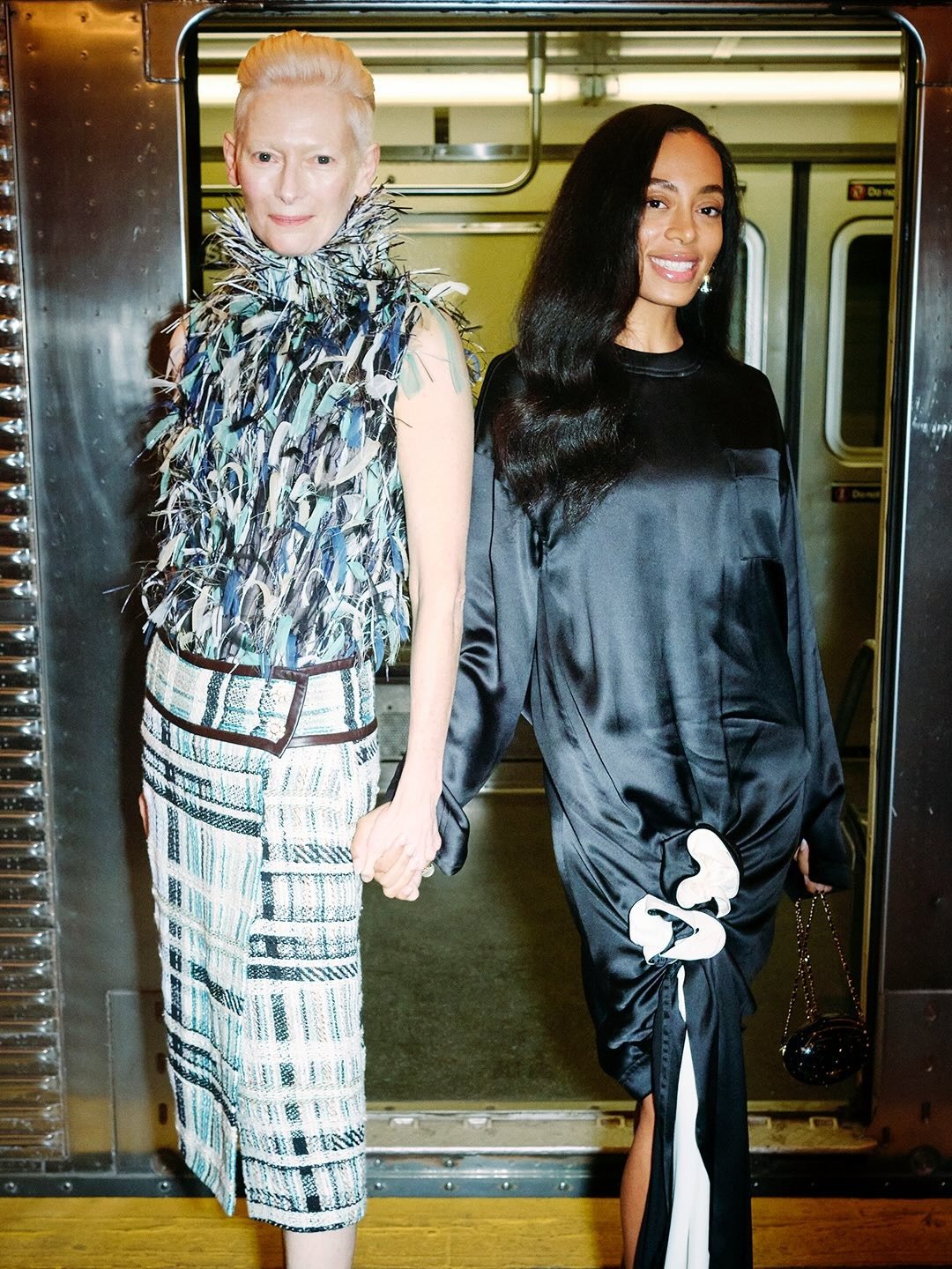 Famosas posam unidas em looks contrastantes: top de penas texturizadas com saia xadrez e vestido preto acetinado com detalhe