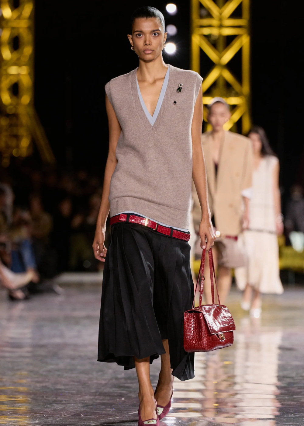 Outono/Inverno 2026: modelo desfila com tricô bege oversized, saia preta fluida e acessórios vinho em clima sofisticado.