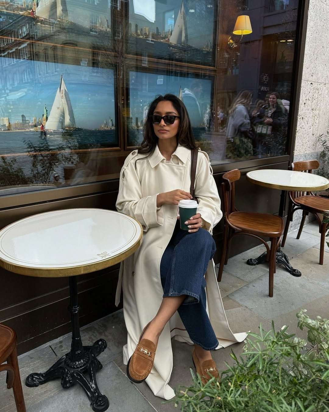 Mocassim feminino com calça jeans e trench coat off-white; modelo posa em café com elegância serena e tons terrosos.