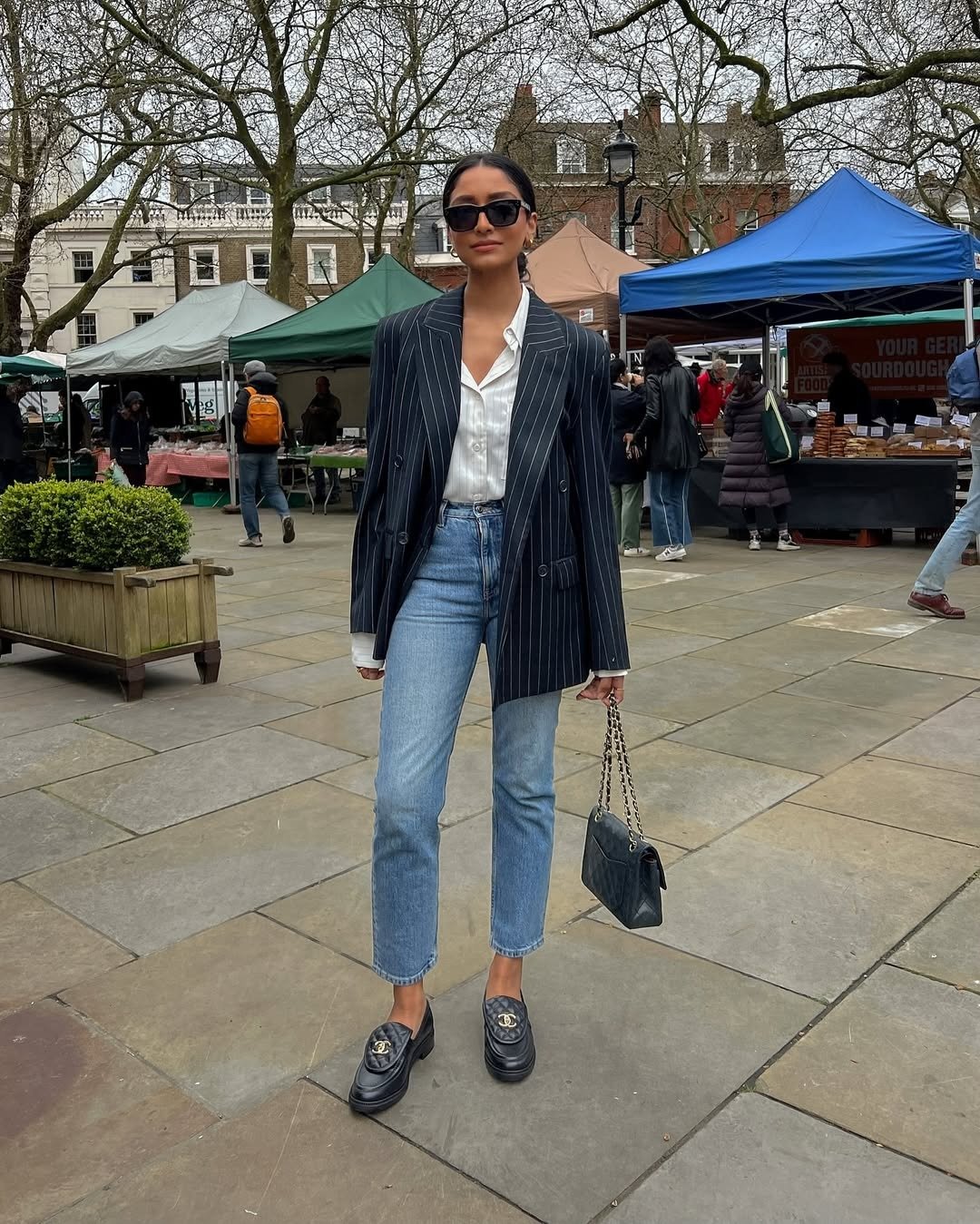 Modelo posa com mocassim feminino com calça jeans, blazer risca de giz e camisa branca em praça urbana elegante.