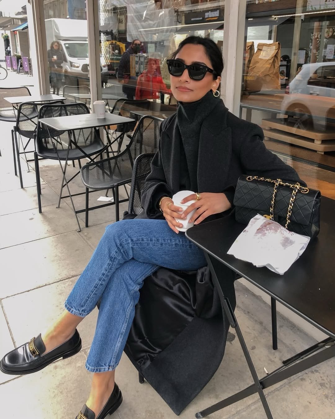 Mocassim feminino com calça jeans: modelo posa em café com casaco preto, tricô canelado e elegância urbana serena.