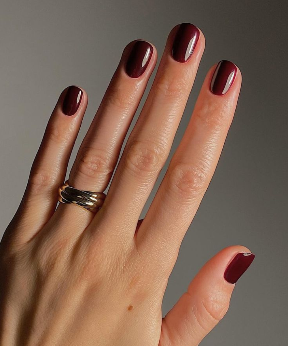 Como fazer o esmalte durar mais com unhas vinho brilhantes e anel dourado sobre fundo cinza, em clima sofisticado.