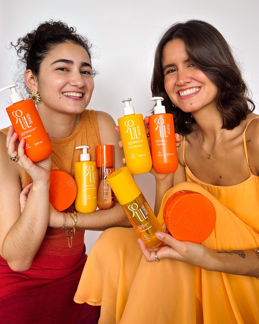 Duas mulheres sorriem segurando produtos hidratantes e perfumados em embalagens laranja e amarelo vibrante, com roupas em