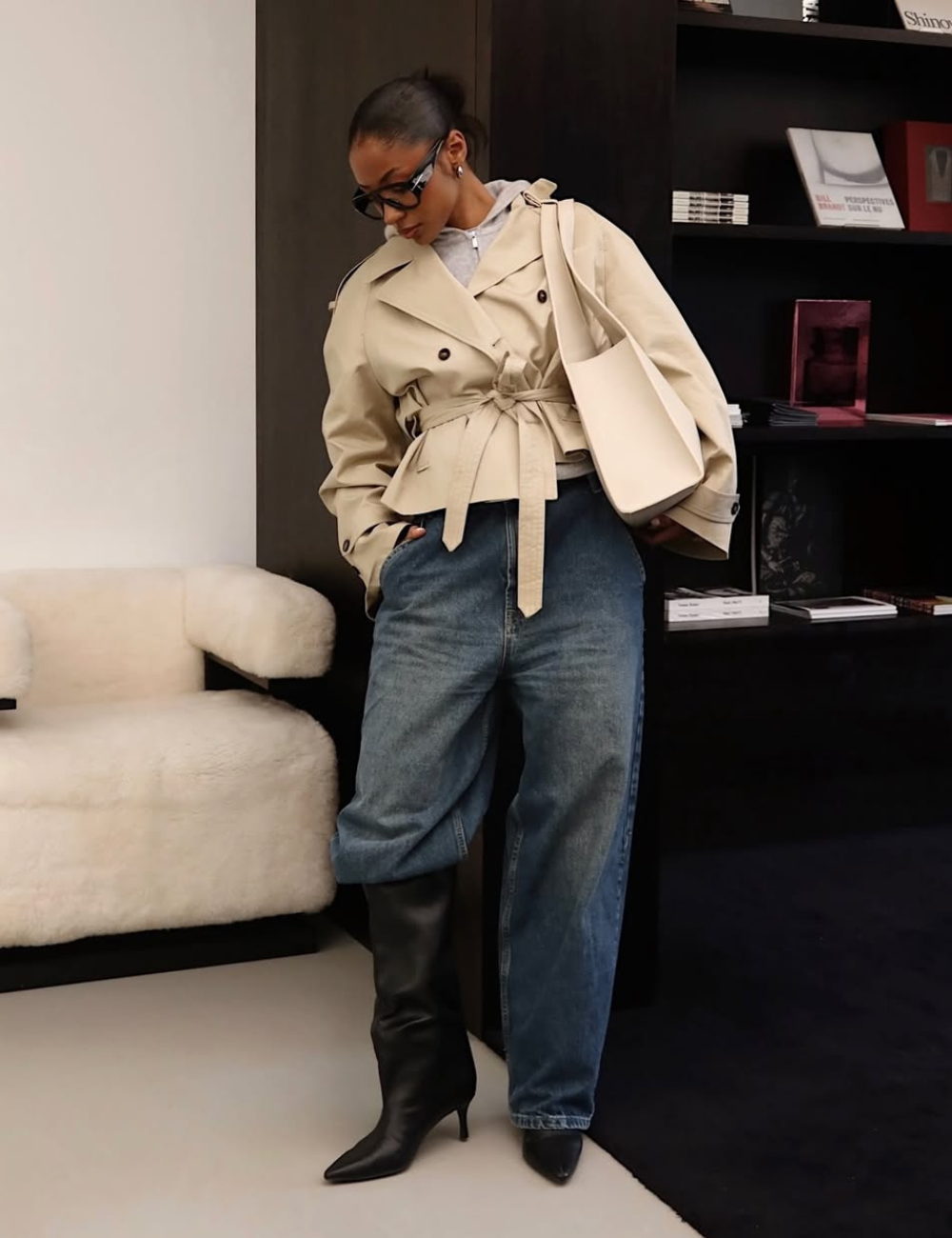 Mulher posa em pé usando bota longa, calças jeans e um trench coat curto.