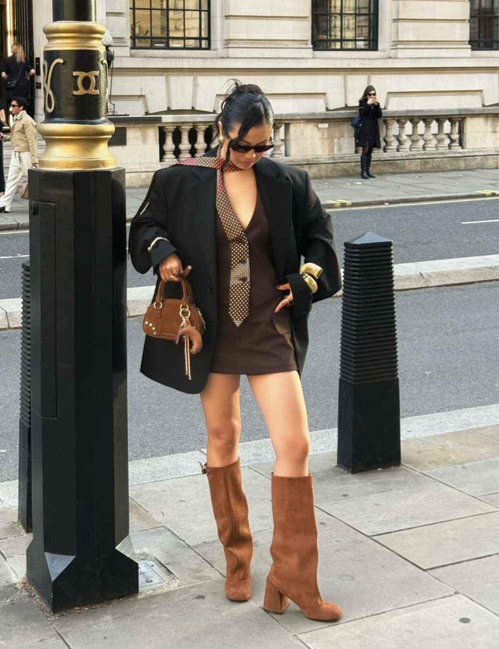 Mulher em pé na rua usando mini vestido, bota longa marrom, blazer oversized preto. Clima urbano e descontraído.