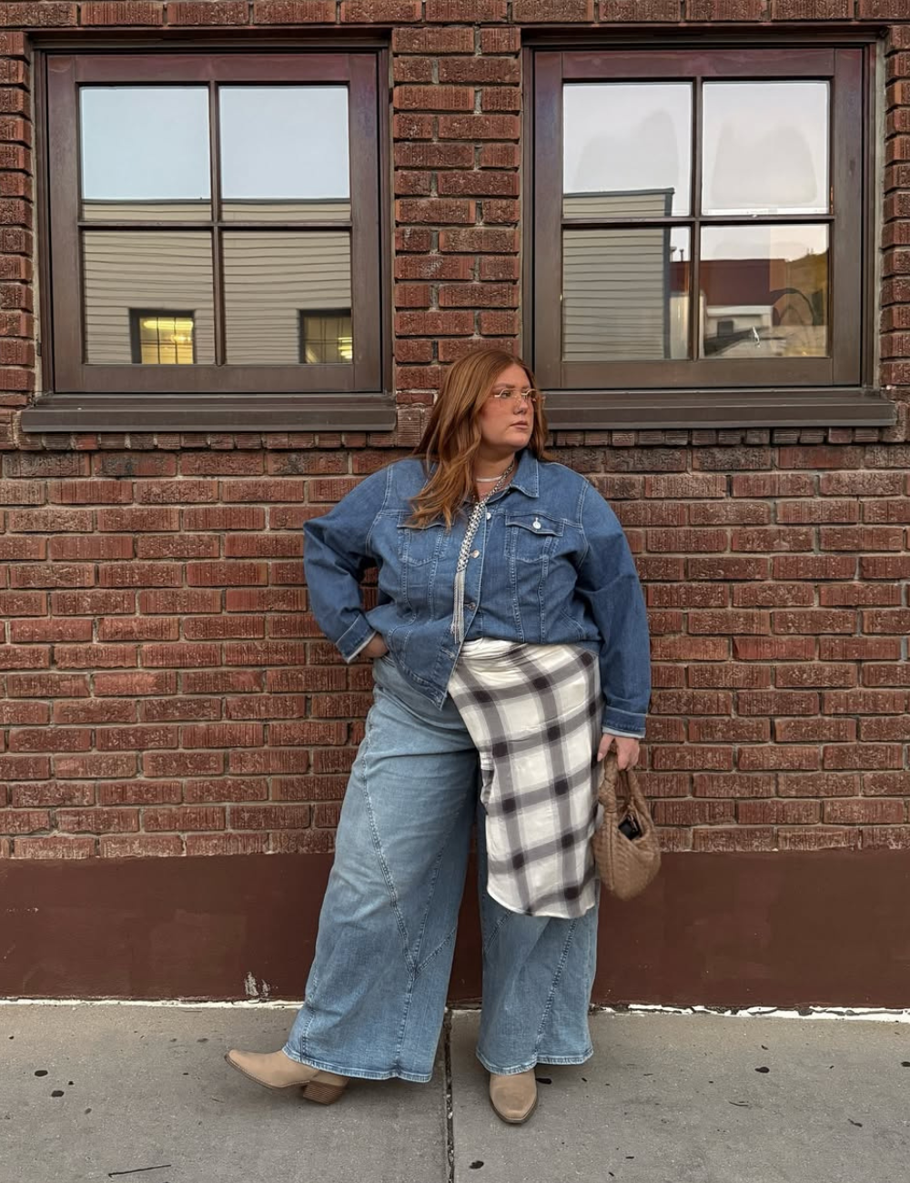 Mulher em pé usando look inteiro jeans com camisa xadrez amarrada na cintura, em frente a uma parede de tijolos.