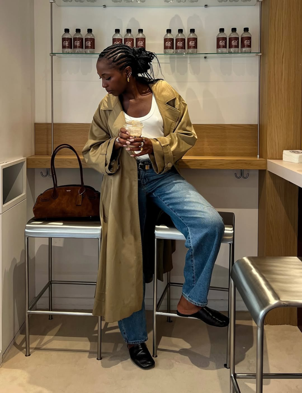 Modelo posa sentada com mule preto de couro, jeans azul, regata branca e trench coat bege, em clima casual e urbano.