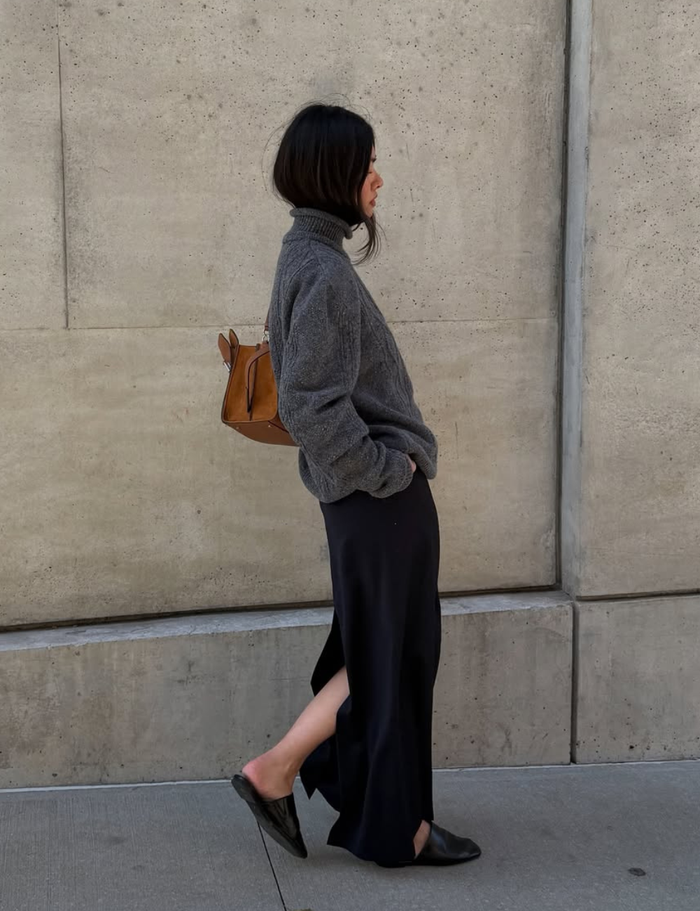 Modelo caminha com postura relaxada usando mule preta de couro, saia longa preta com fenda e suéter cinza oversized.