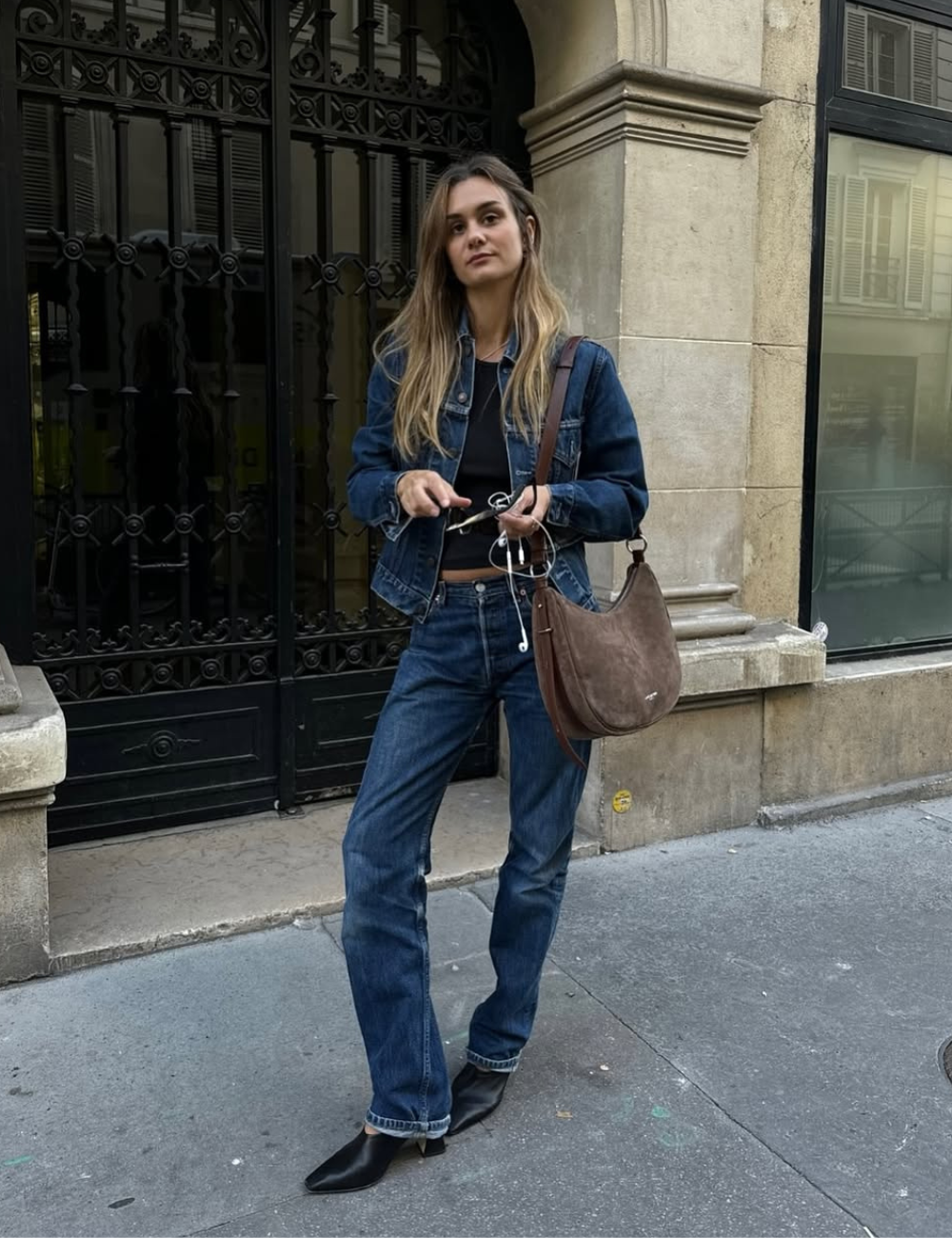 Modelo posa em look casual com jaqueta e jeans azul escuro, mule preto de bico fino em couro, bolsa marrom e top preto.