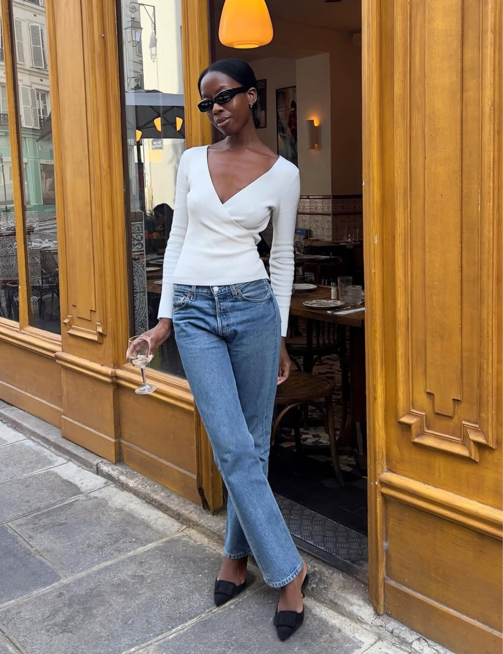 Modelo posa descontraída com blusa branca de manga longa e decote V, jeans azul reto e mule preta de bico fino.