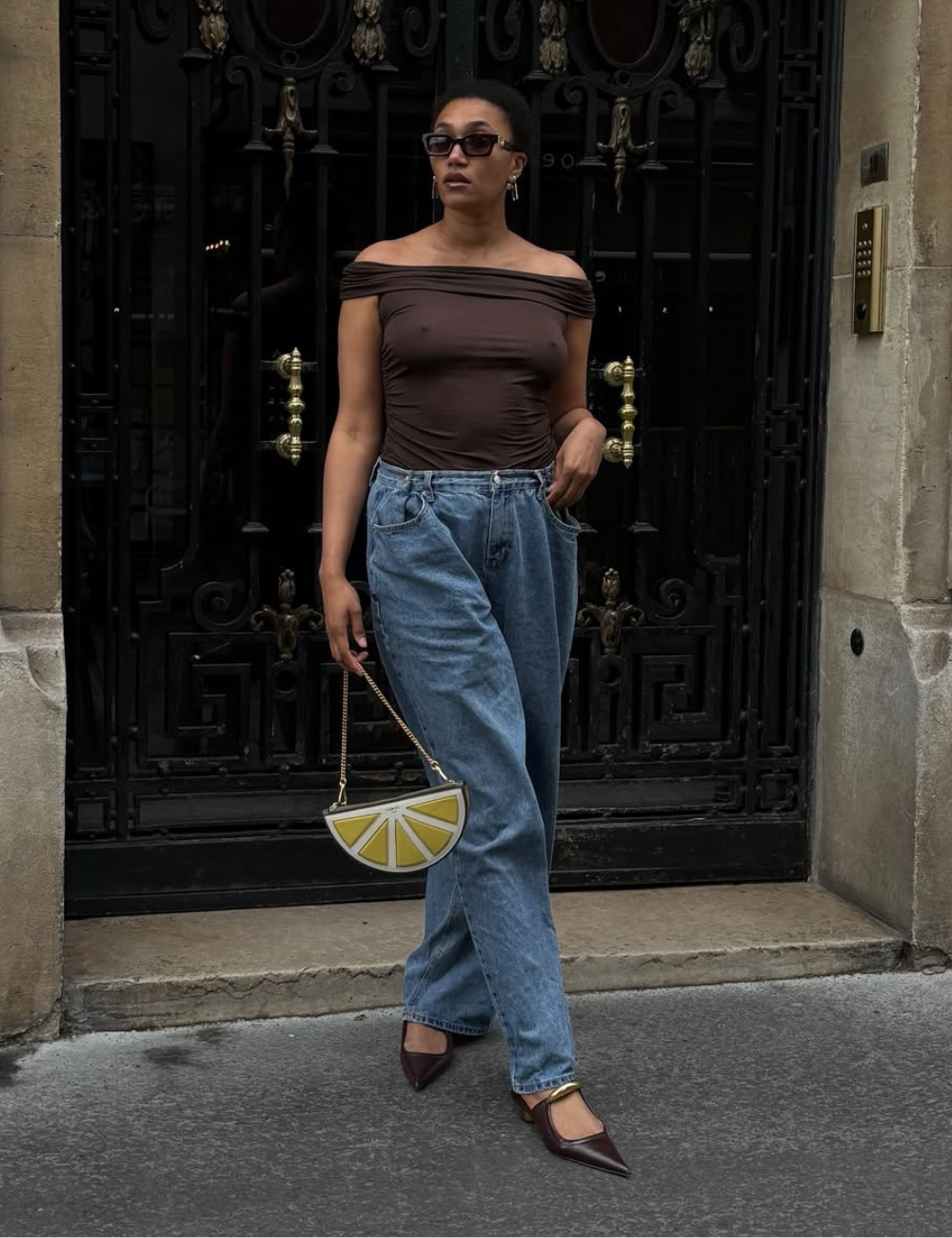 Modelo posa com blusa ombro a ombro marrom, jeans largo azul e mule de couro marrom com bico fino, mostrando como usar mule.