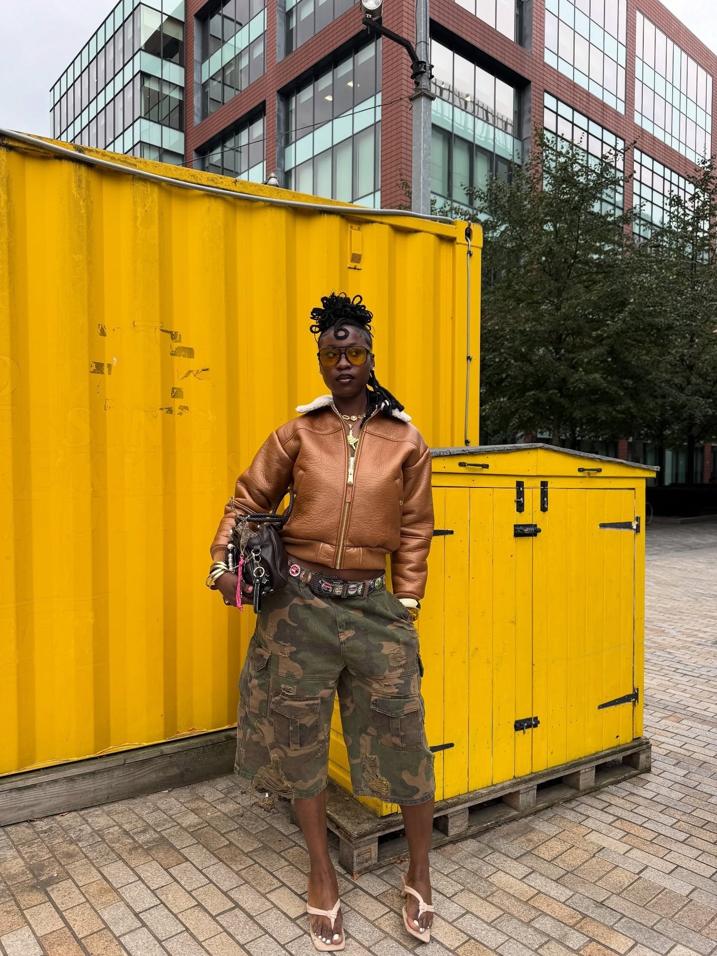 Modelo posa com jaqueta marrom, bermuda cargo camuflada e sandália nude, uma das cores de sapato que combinam com tudo.