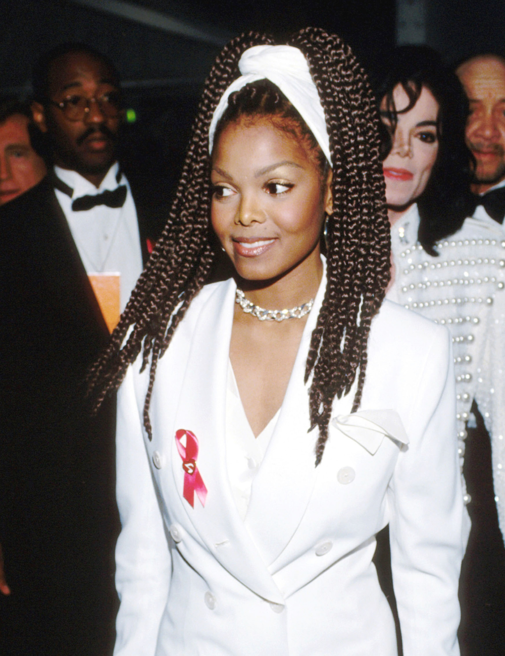 Cortes de cabelo dos anos 90 em foco: modelo posa com tranças longas, turbante e blazer branco, em clima elegante.