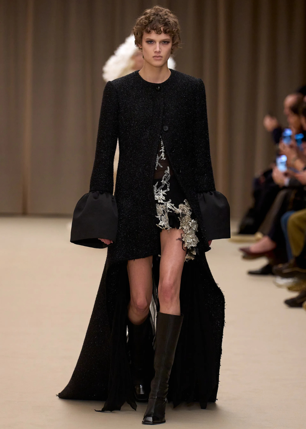 Outono/Inverno 2026: modelo desfila vestido preto fluido com transparência e babados, em passarela enevoada, clima dramático.