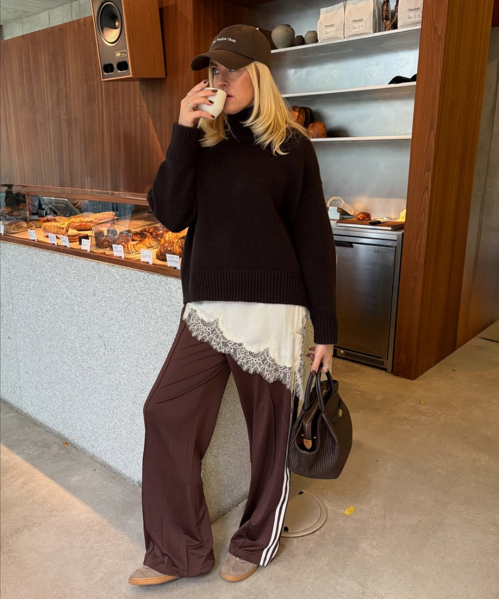 Modelo posa em cafeteria com calça pantalona marrom, tênis bege, tricô escuro e renda branca.