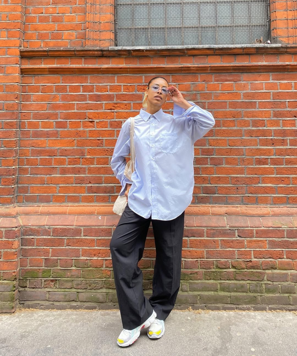 Modelo posa com calça pantalona preta e tênis brancos, camisa azul ampla e bolsa clara diante de parede de tijolos.