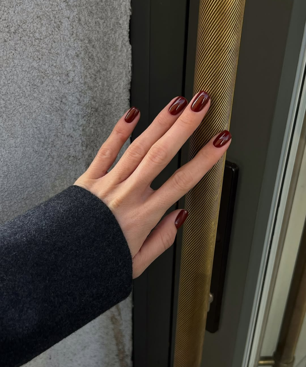 Esmaltes para o outono em tom vinho brilhante destacam mão alongada com casaco escuro sobre puxador dourado elegante.