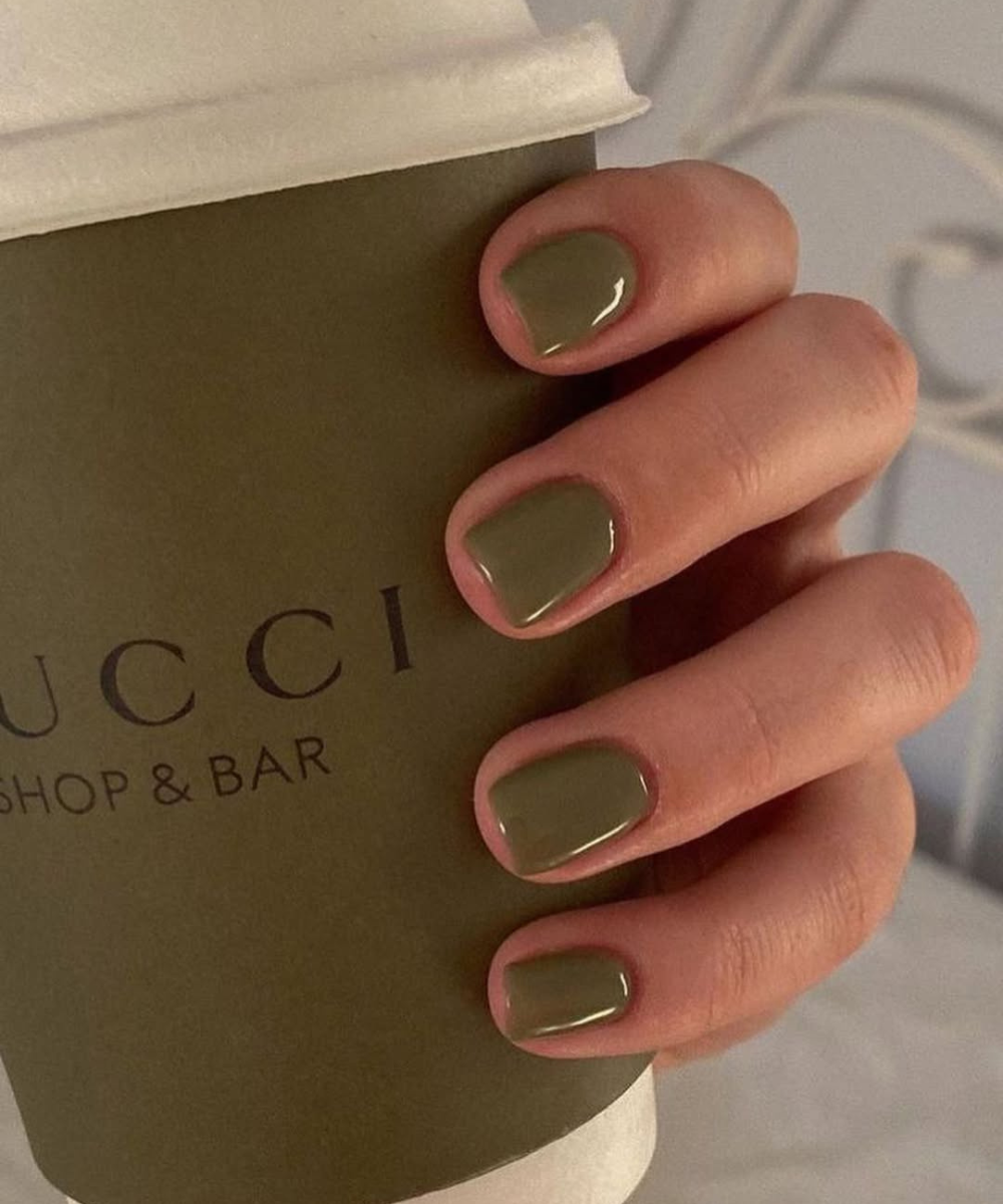 Esmaltes para o outono em verde-oliva cremoso brilham nas unhas curtas enquanto a mão segura copo neutro.