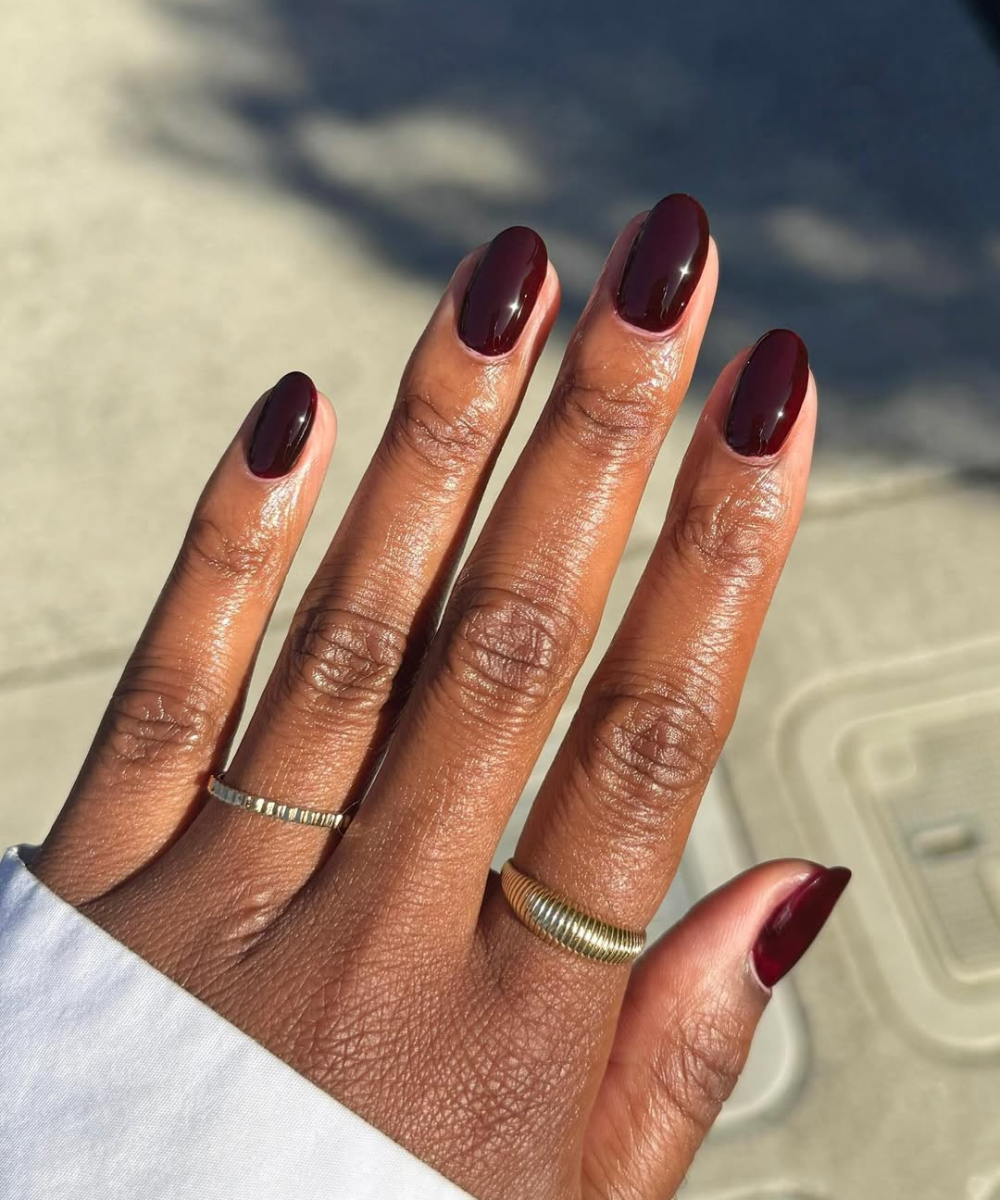 Esmaltes para o outono em vinho escuro brilham nas unhas amendoadas, com anéis dourado e prateado sob luz quente.