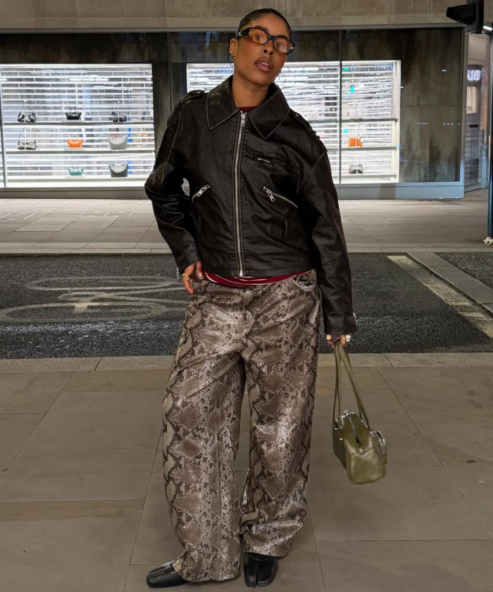 Modelo posa com calça resinada animal print ampla, jaqueta preta e bolsa verde em cenário urbano de atitude sofisticada