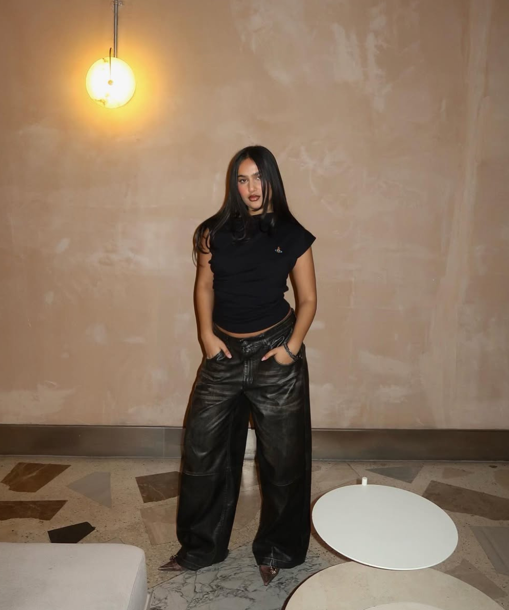 Modelo posa com calça resinada preta ampla e blusa ajustada preta em ambiente bege de luz quente e clima sofisticado.