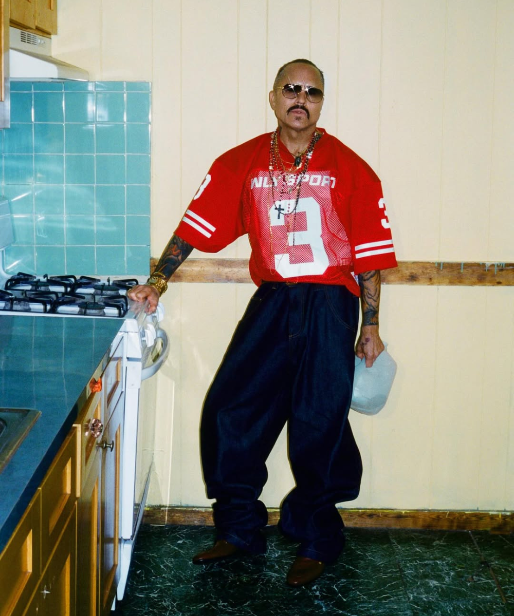 Willy Chavarria posa na cozinha com jersey vermelho oversized e calça ampla azul-marinho;